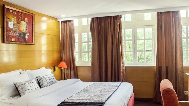 A hotel room with a bed, a chair, a window with curtains, and a painting on the wall. - HOTELS DAFFODILS (New Delhi)