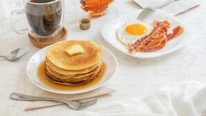 A breakfast setting with pancakes, a fried egg, bacon, a glass of coffee, and a bottle of syrup. - HOTELS DAFFODILS (New Delhi)