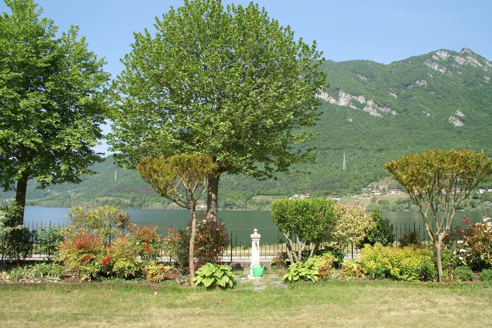 Un parque tranquilo junto al lago, con abundante vegetación, una fuente blanca y montañas al fondo.