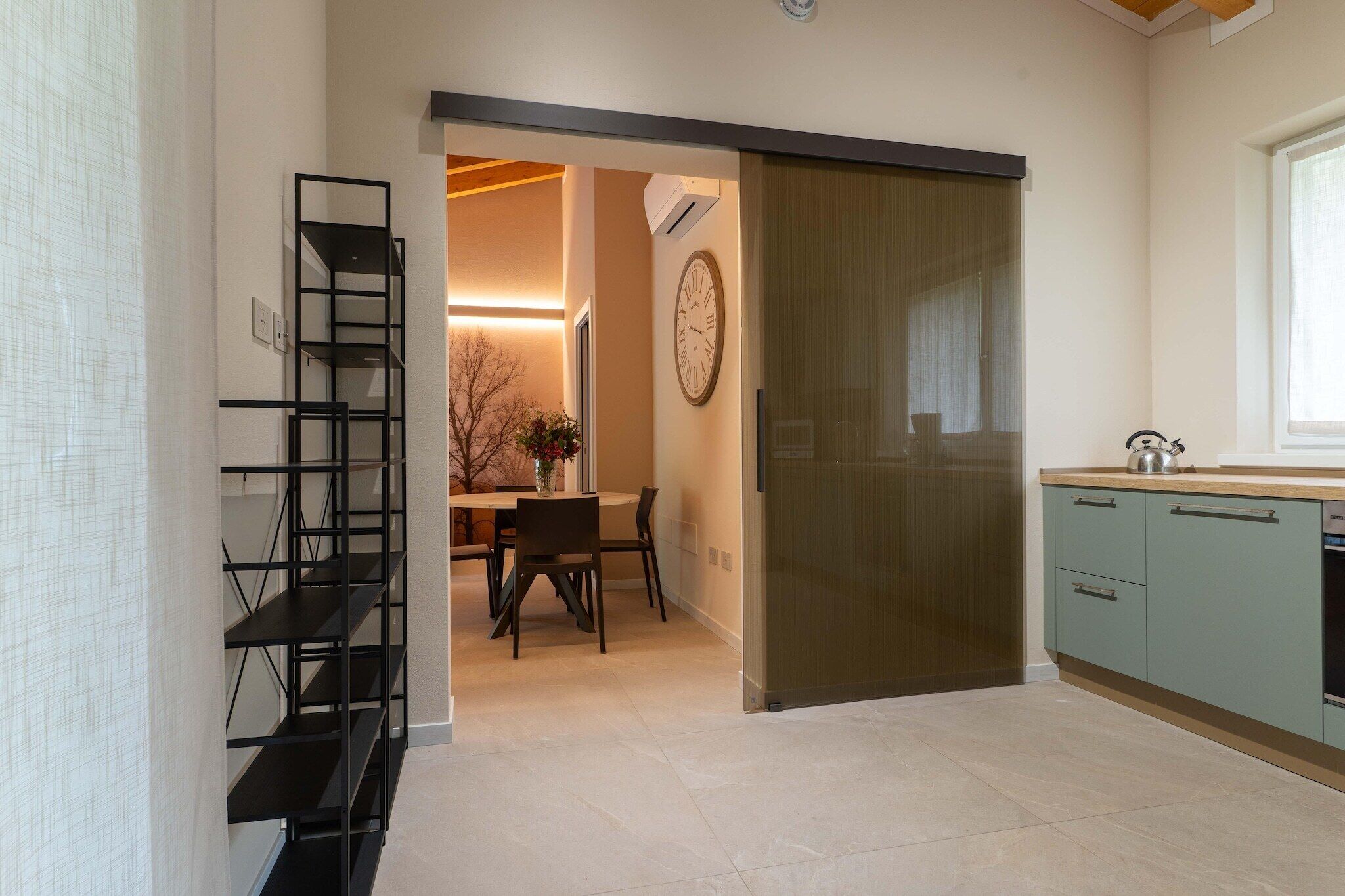 Una cocina moderna con puerta corrediza, un comedor con mesa y sillas, y una estantería metálica negra.