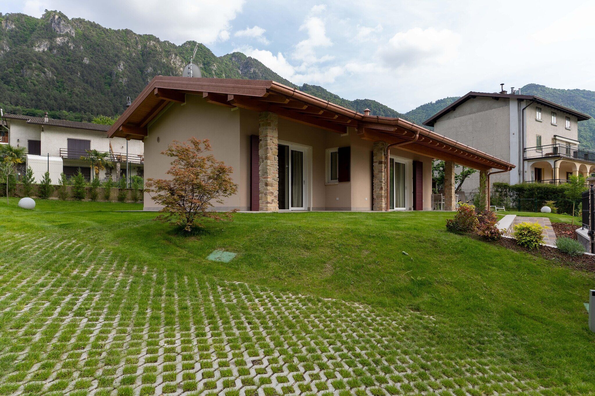 A house with a stone facade, a wooden roof, and a paved lawn with a small tree.