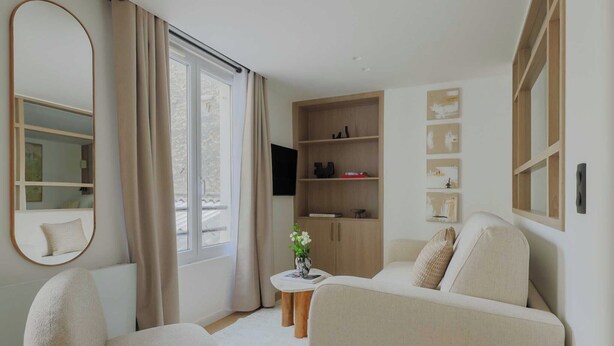 Un salon moderne avec un canapé, un fauteuil et une petite table sur laquelle est posé un vase. Un grand miroir orne le mur, et une étagère en bois est décorée de divers objets.