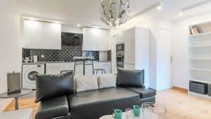 A modern living room with a black leather sofa, a glass coffee table, and a kitchen area in the background. - Comfortable Apartment - 1BR/2P - Arts et Métiers (Paris)
