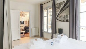 A hotel room with a bed, a radiator, a window with curtains, a cityscape view, and a door leading to another room. - Comfortable Apartment - 1BR/2P - Arts et Métiers (Paris)
