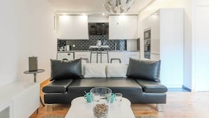 A modern living room with a black leather sofa, a white coffee table with decorative items, and a kitchen in the background with white cabinets and a black backsplash. - Comfortable Apartment - 1BR/2P - Arts et Métiers (Paris)