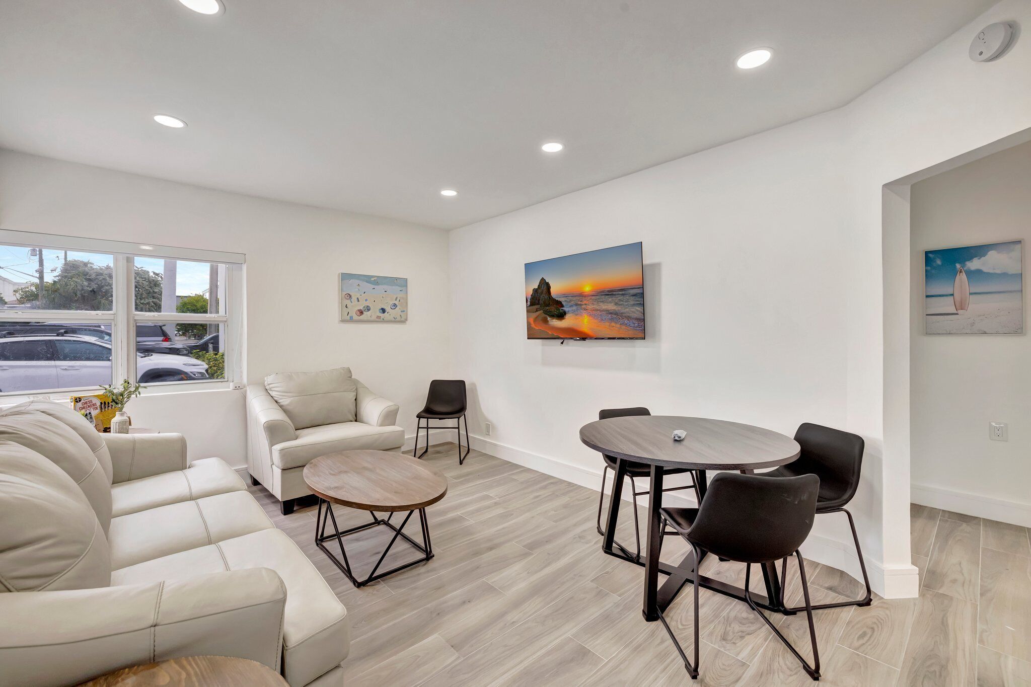 A modern living room with a sofa, chairs, a coffee table, and a dining table with chairs. There are paintings on the walls and a window with a view of parked cars outside.