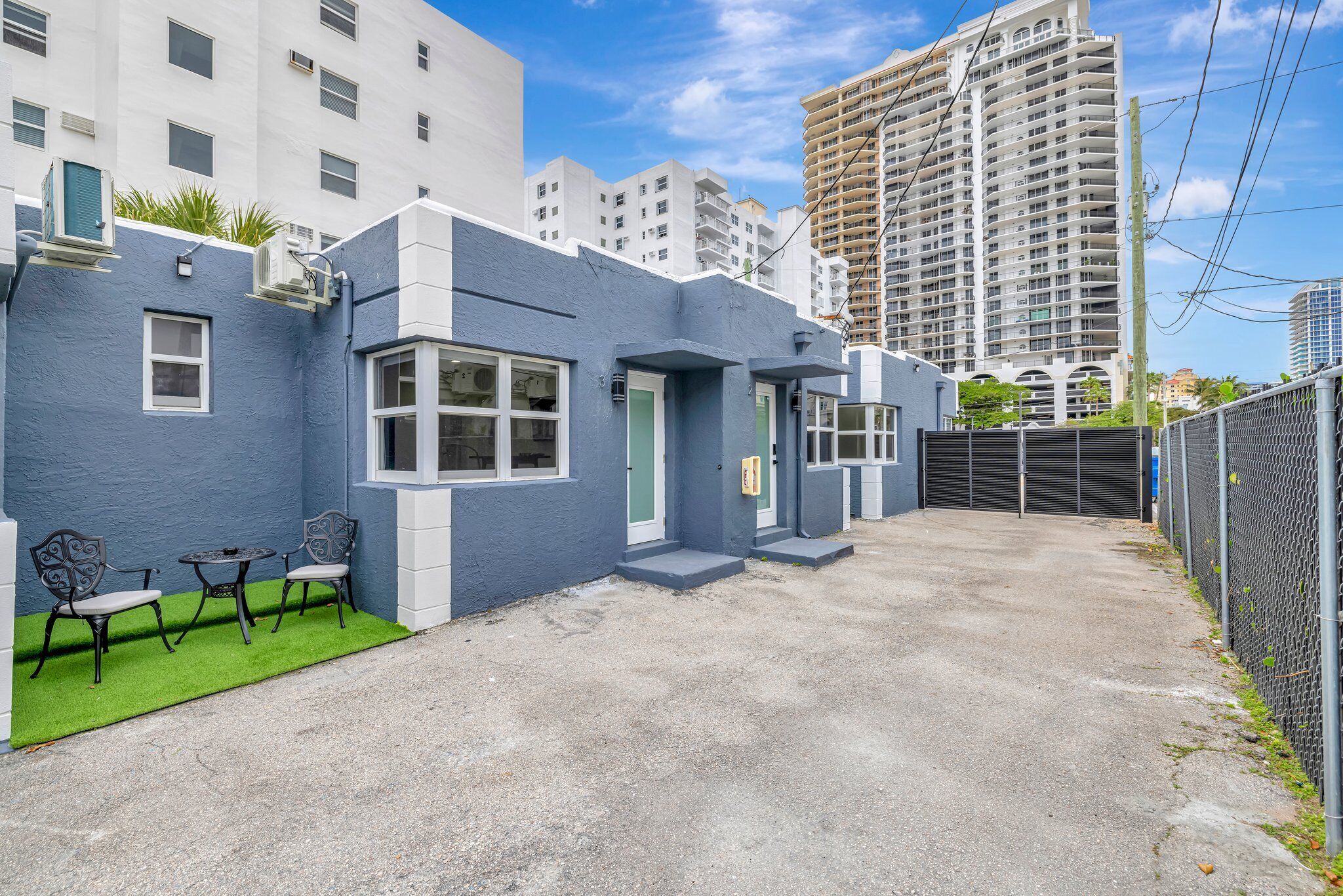 A modern building with a blue exterior, a small patio with chairs, and a gated entrance.