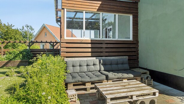 Un espace extérieur moderne, aménagé avec un banc en bois et des coussins, une table en bois et une grande fenêtre donnant sur un jardin.