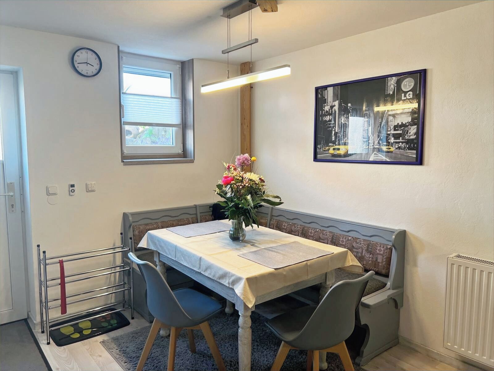 A dining area with a table, chairs, a vase of flowers, a clock, and a framed picture on the wall.