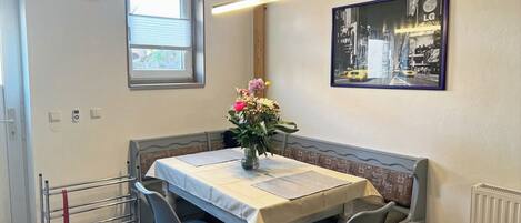 A dining area with a table, chairs, a vase of flowers, a clock, and a framed picture on the wall.