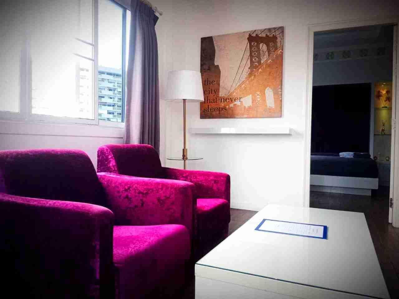 Une chambre d’hôtel avec une grande fenêtre, une vue sur la ville, un lit, un bureau et deux fauteuils roses.