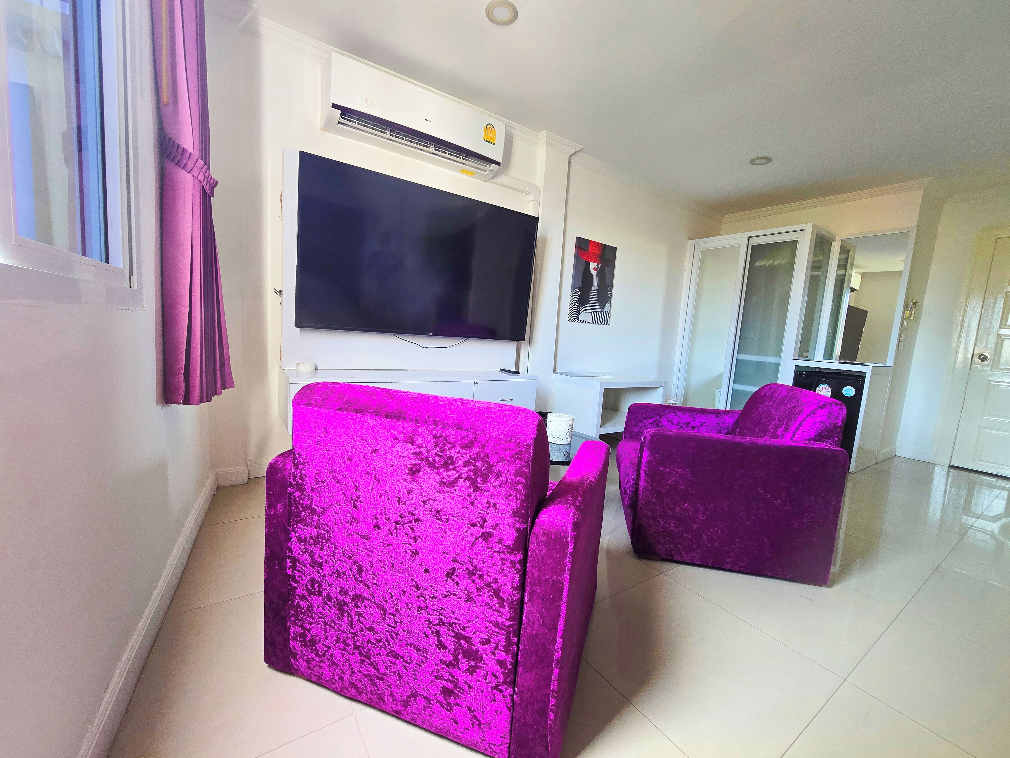 A living room with a flat-screen TV, purple velvet sofas, a white entertainment unit, and a large mirror.