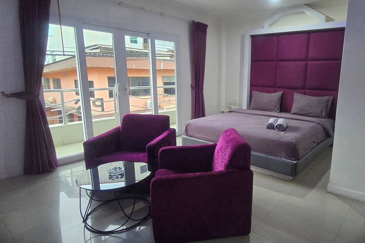 A hotel room with a bed, two purple armchairs, a small table, and a view of a building through a glass door.