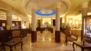 Un vestíbulo con mostrador de recepción, varias sillas, una columna central iluminada en azul y suelo de baldosas.