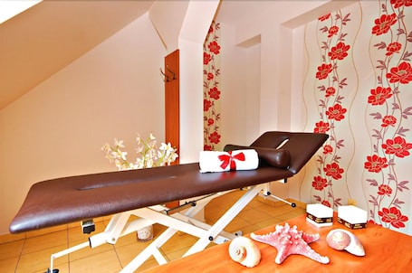 A medical examination room with a treatment bed, a small table with a towel and a bottle, and a decorative wall with floral wallpaper.