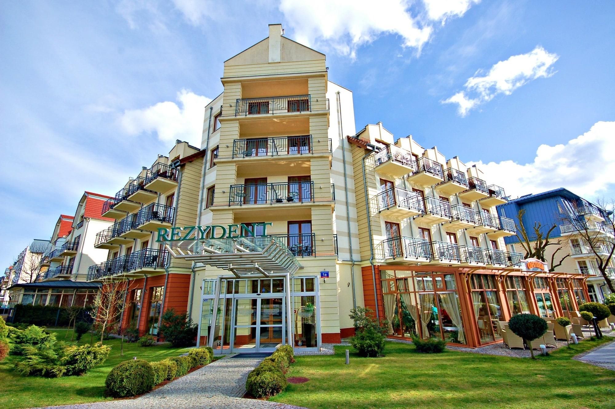 Un hotel de varios pisos con una entrada central, balcones en cada planta y un jardín bien cuidado.
