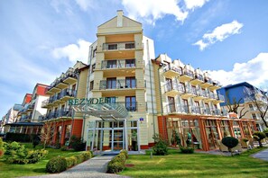 Um hotel com vários andares, entrada central, varandas em cada pavimento e um jardim bem cuidado.