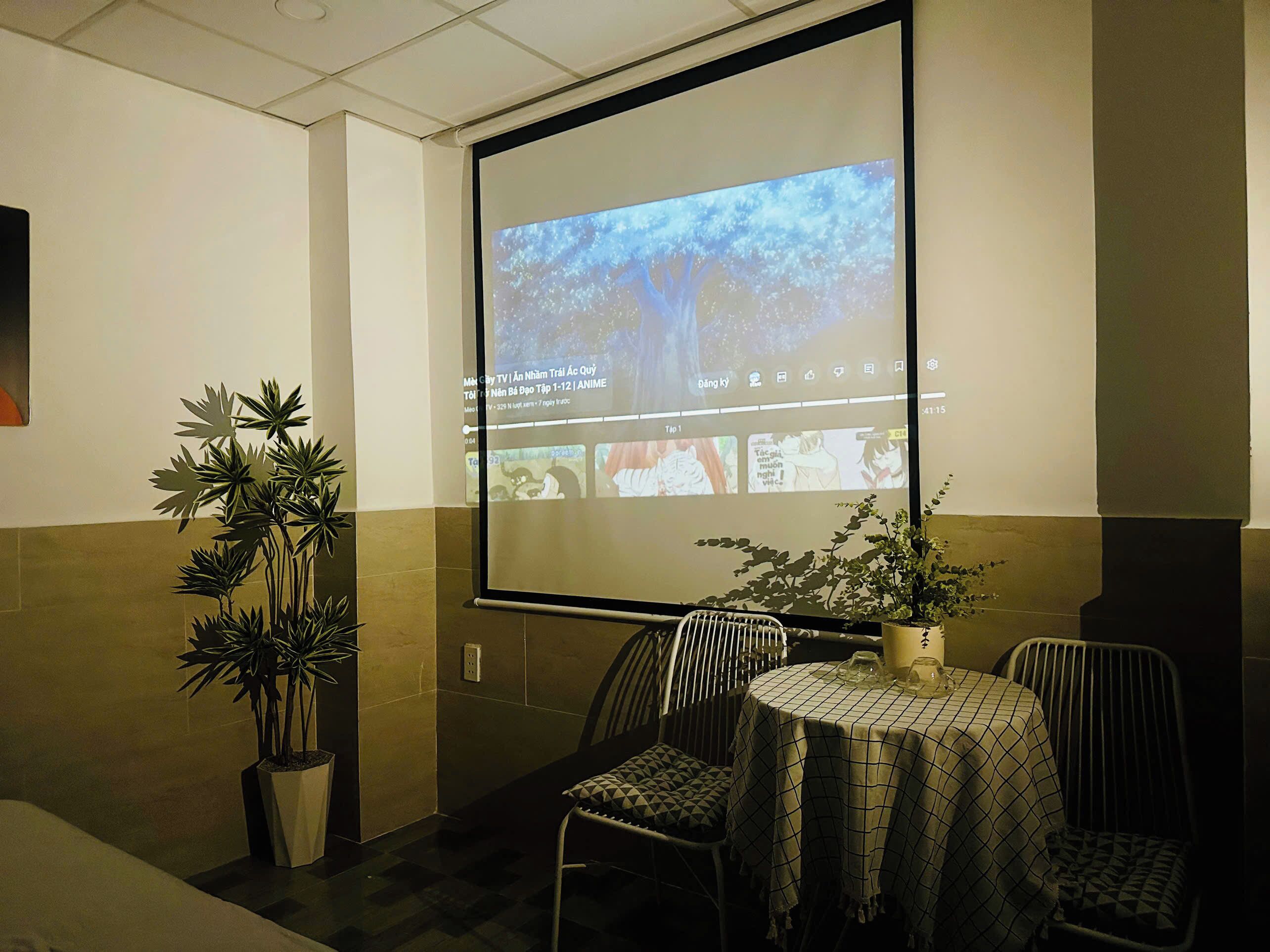 A conference room with a projection screen displaying an anime, a potted plant, and a table with chairs.