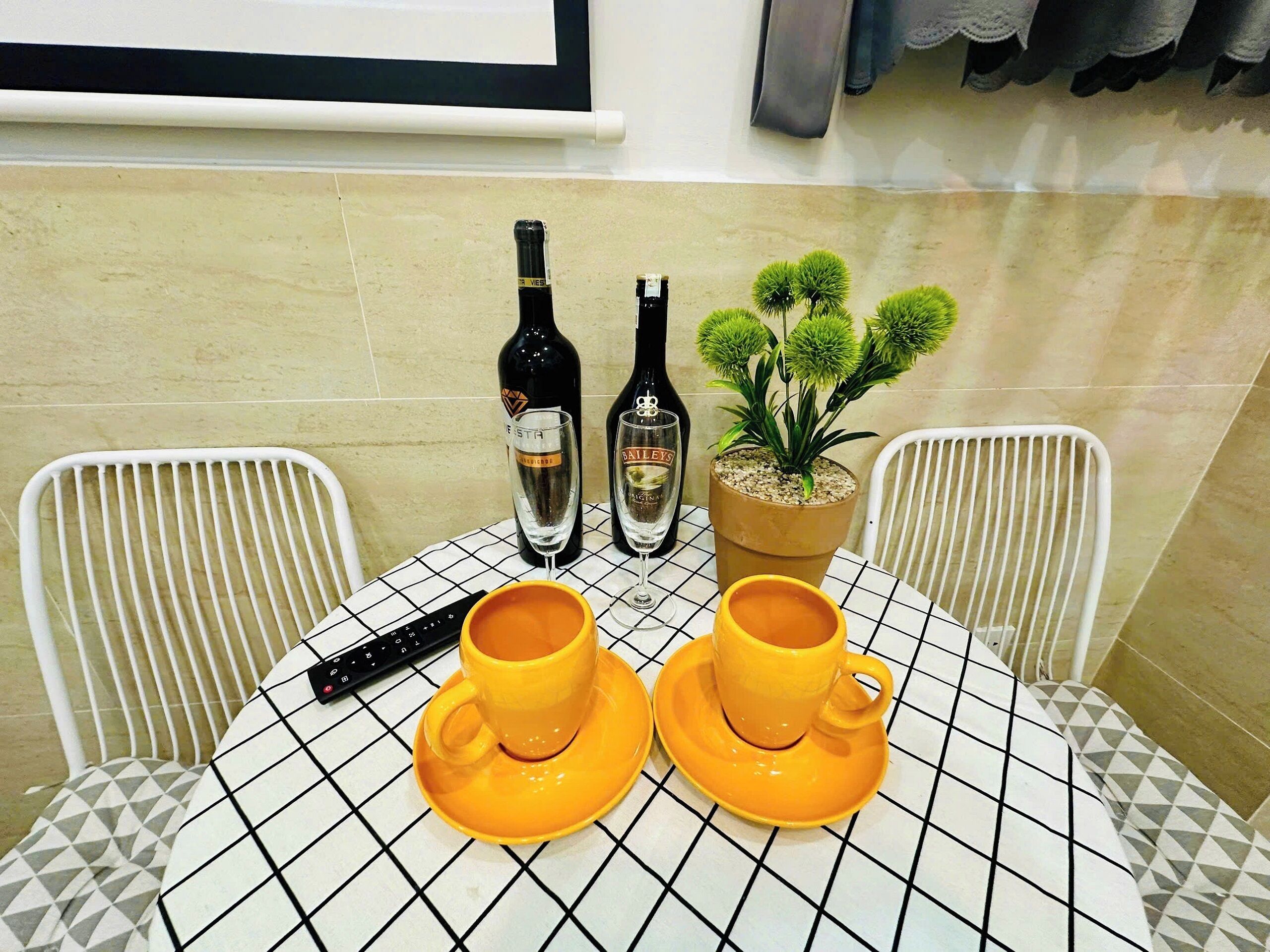 Una mesa con dos tazas amarillas y dos botellas de vino, una planta en maceta y un control remoto.