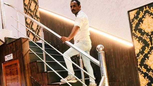 A man ascending a staircase with a metal handrail, leading to a room with a bed and a wooden door labeled "TOILET". - The Nest Ayodhya (Faizabad)