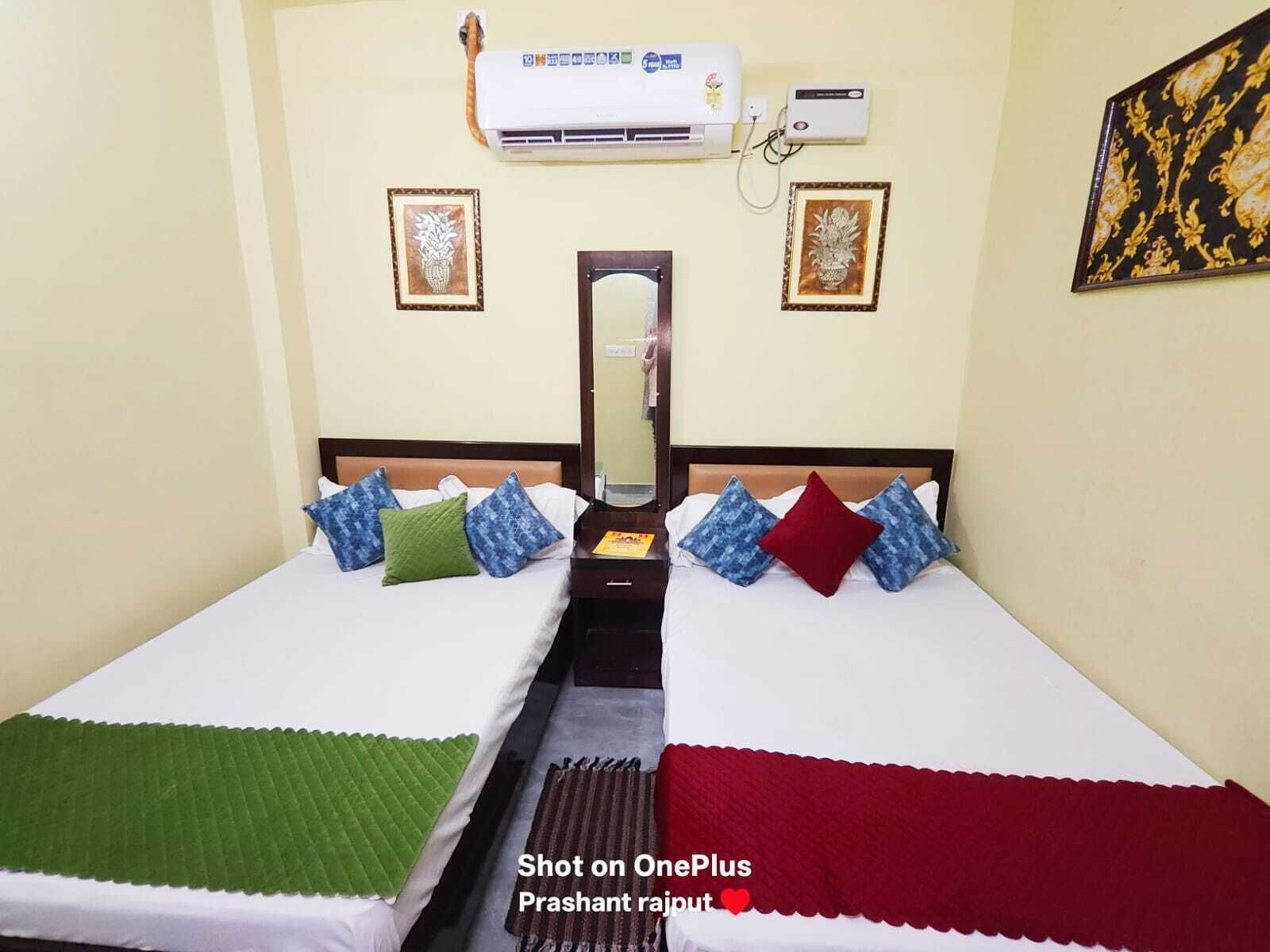 Habitación de hotel con dos camas, cada una con un edredón de color diferente, un espejo y dos unidades de aire acondicionado fijadas a la pared.