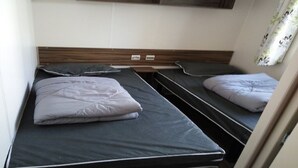 A compact room with two beds, a wooden headboard, and a window with a floral curtain. - SABLES DU MIDI LOCATION CLIM 3CH 2SDB CAMPING VOS VACANCES (Valras-Plage)