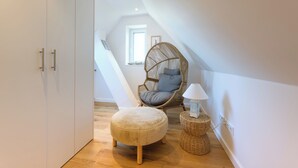 Un dormitorio tipo loft acogedor con una silla de ratán, un pequeño reposapiés y una lámpara sobre una mesita de noche tejida.