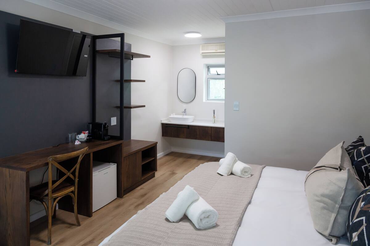 Een compacte hotelkamer met een bed, een bureau met stoel, een kleine koelkast, een aan de muur bevestigde televisie, een spiegel en een raam.