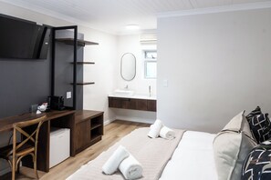 Une chambre d’hôtel moderne équipée d’un bureau, d’un petit réfrigérateur, d’un lit avec du linge de maison blanc et d’une salle de bain avec un lavabo et un miroir.