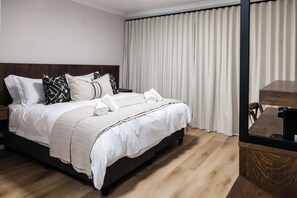 Un lit bien fait, avec du linge de lit blanc et de nombreux oreillers, une tête de lit en bois, un plancher en bois et une grande fenêtre avec des rideaux.