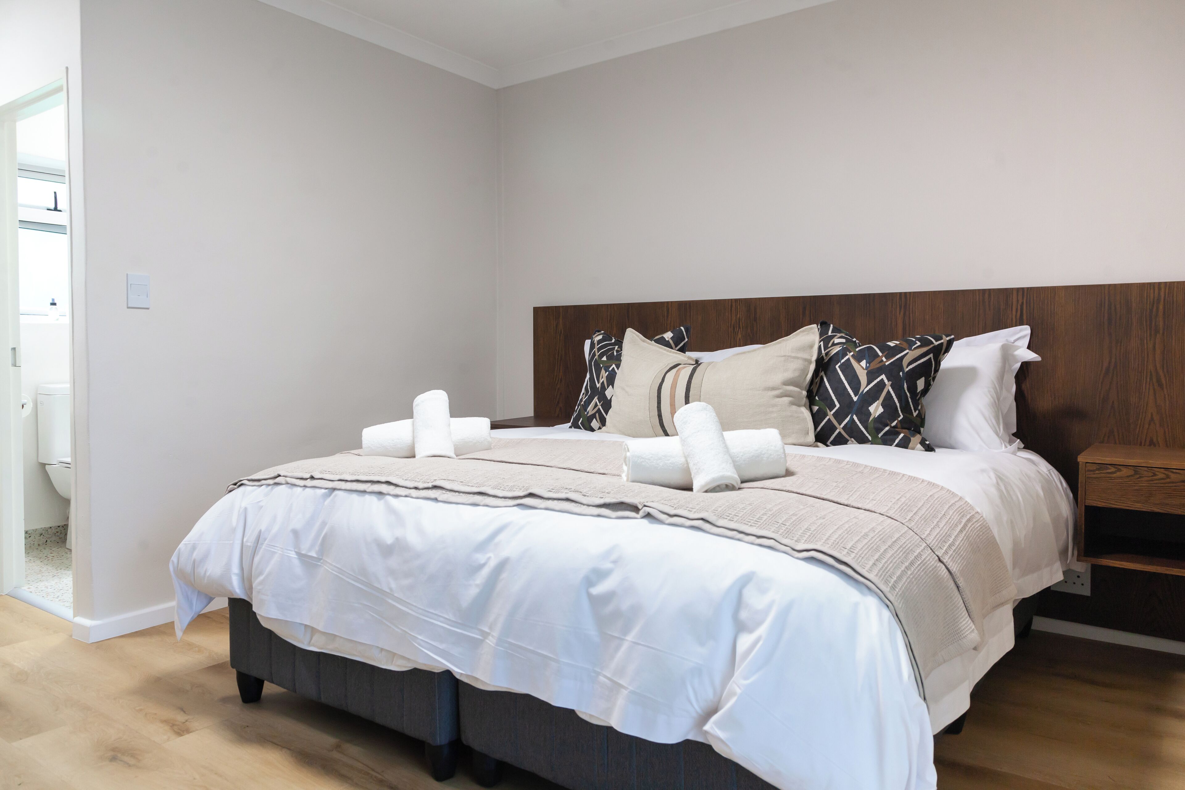 A neatly made bed with a wooden headboard, a bedside table, and a view of a bathroom with a toilet and sink.