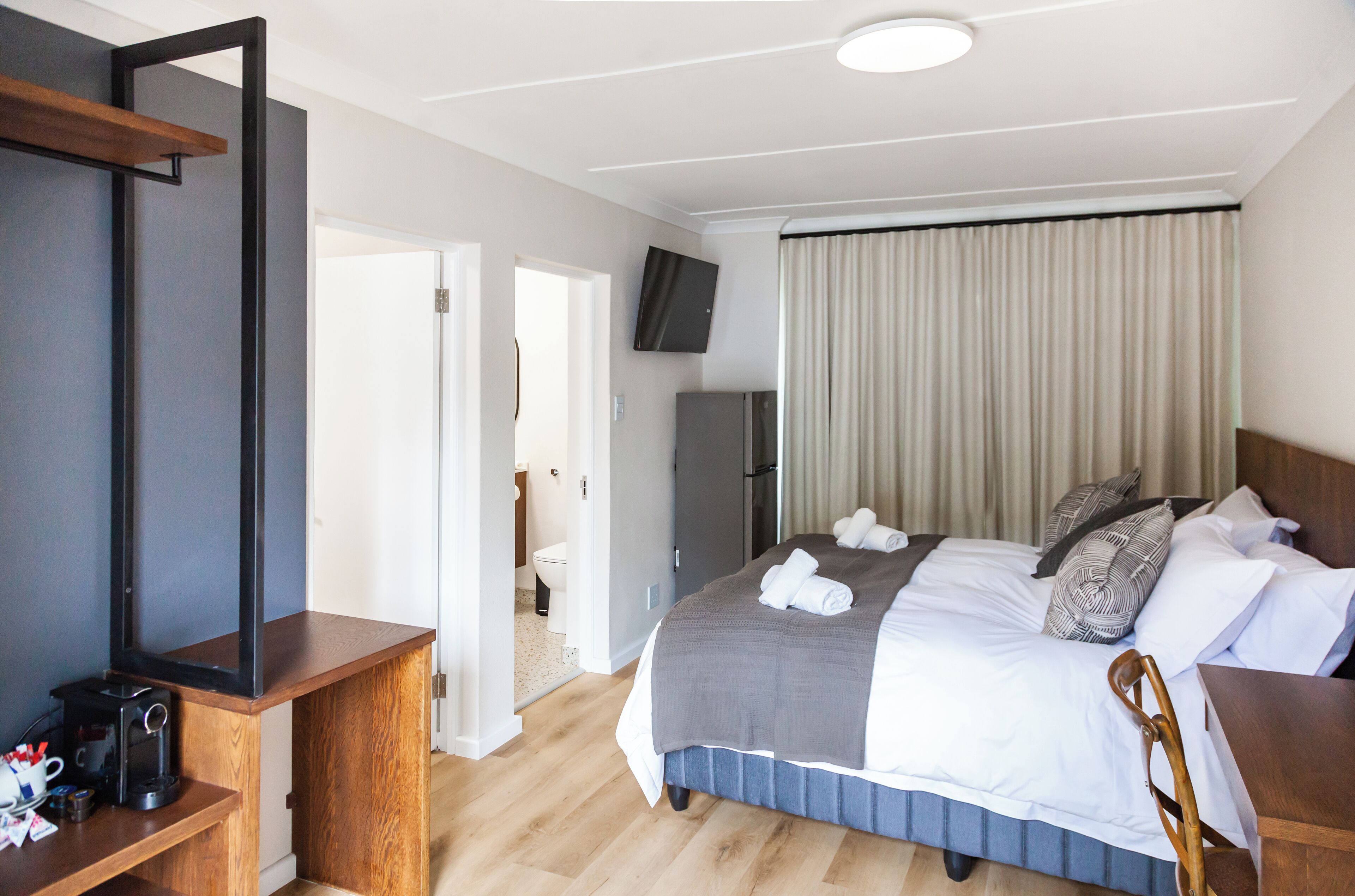 A hotel room with a bed, a TV mounted on the wall, a desk with a coffee machine, and a door leading to the bathroom.