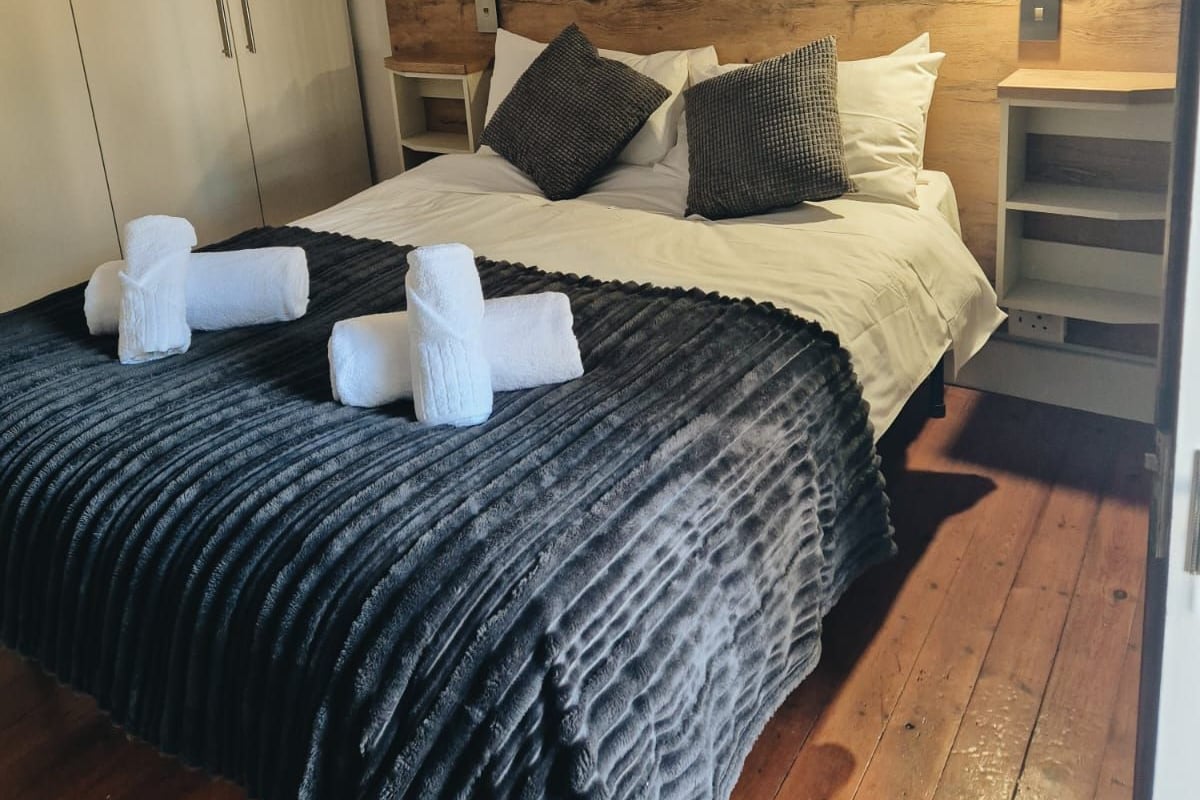Un dormitorio con cabecera de madera, cama con ropa blanca y una manta negra texturizada. Hay dos toallas blancas sobre la cama.