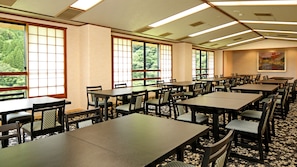 Un salón de conferencias con varias mesas y sillas, ventanas amplias que dan a zonas verdes e iluminación empotrada en el techo.