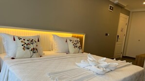 A neatly made bed with white linens and decorative pillows, a bedside table, and a door in the background. - Ege Vadi Otel (Ayvalik)