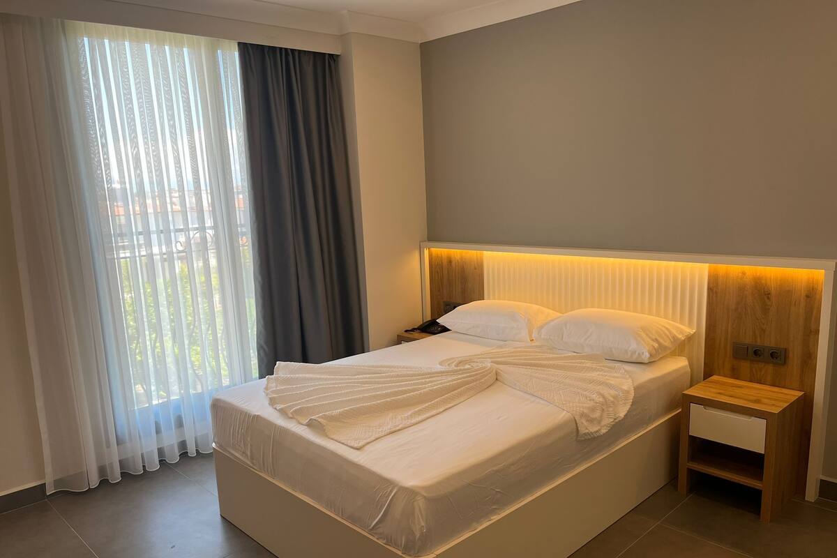 A hotel room with a large bed, a bedside table with a lamp, and a view of greenery through the window.