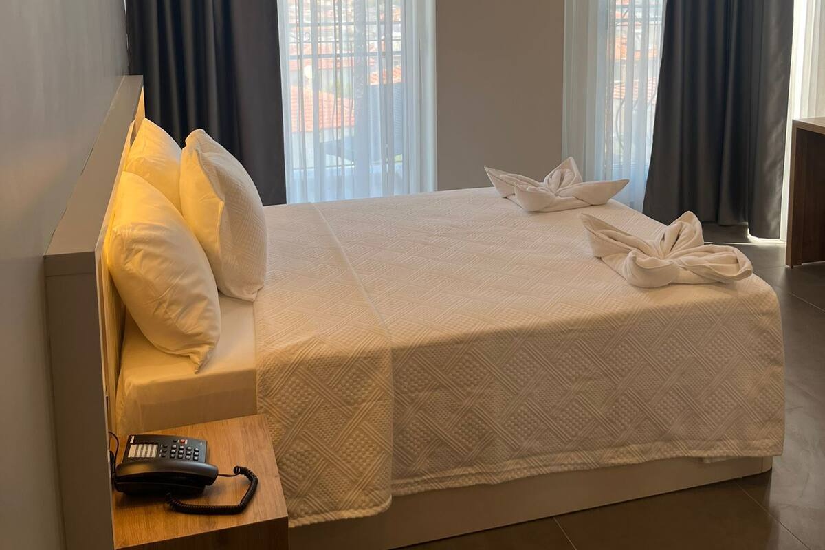A hotel room with a large bed, a bedside telephone, and a view of buildings through the windows.