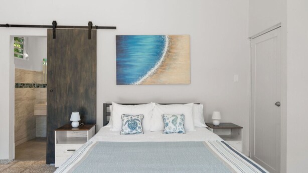 A bedroom with a bed, two nightstands, a wooden sliding door, and a beach-themed painting on the wall. - Dowtown Studio Walkable to Town and Beach (Rincon)