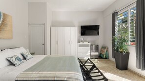 Une chambre à coucher comprenant un lit, une armoire, une télévision, un réfrigérateur, une plante et une fenêtre avec des rideaux.