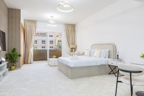 A modern bedroom with a large bed, a small bedside table, a chair, a desk, and a balcony with curtains.