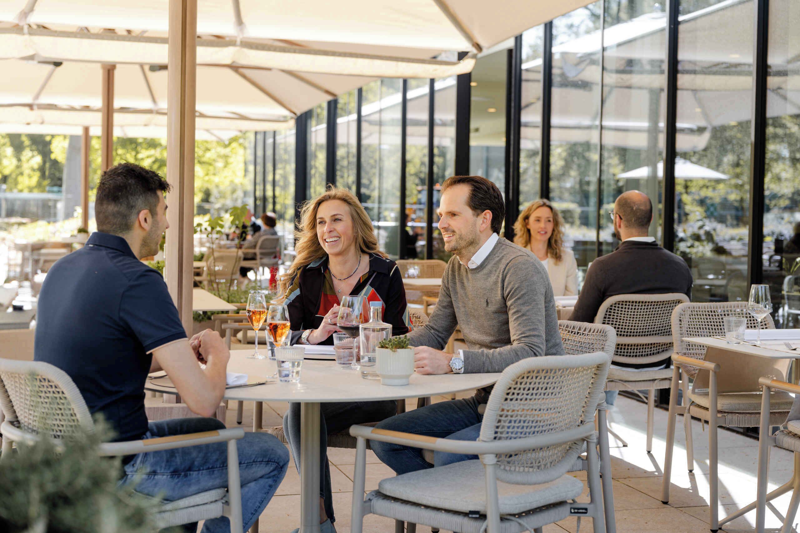 An outdoor dining area with round tables and wicker chairs, large windows, and a view of greenery.