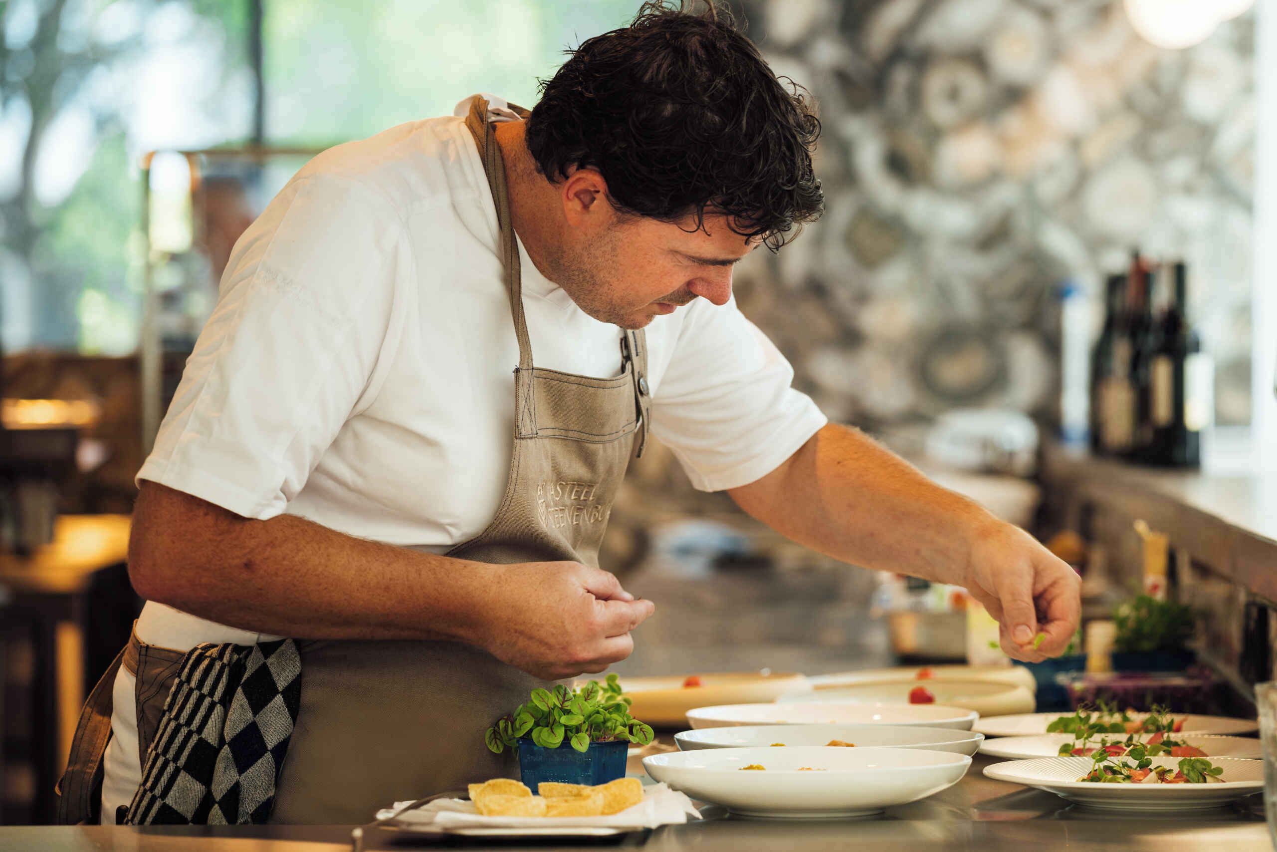 Un chef con camisa y delantal blanco prepara comida en una barra, con platos de comida y una pequeña planta encima.