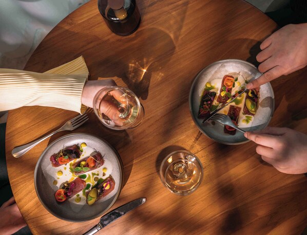 Una mesa de madera redonda con tres platos de comida, una copa de vino y cubiertos.