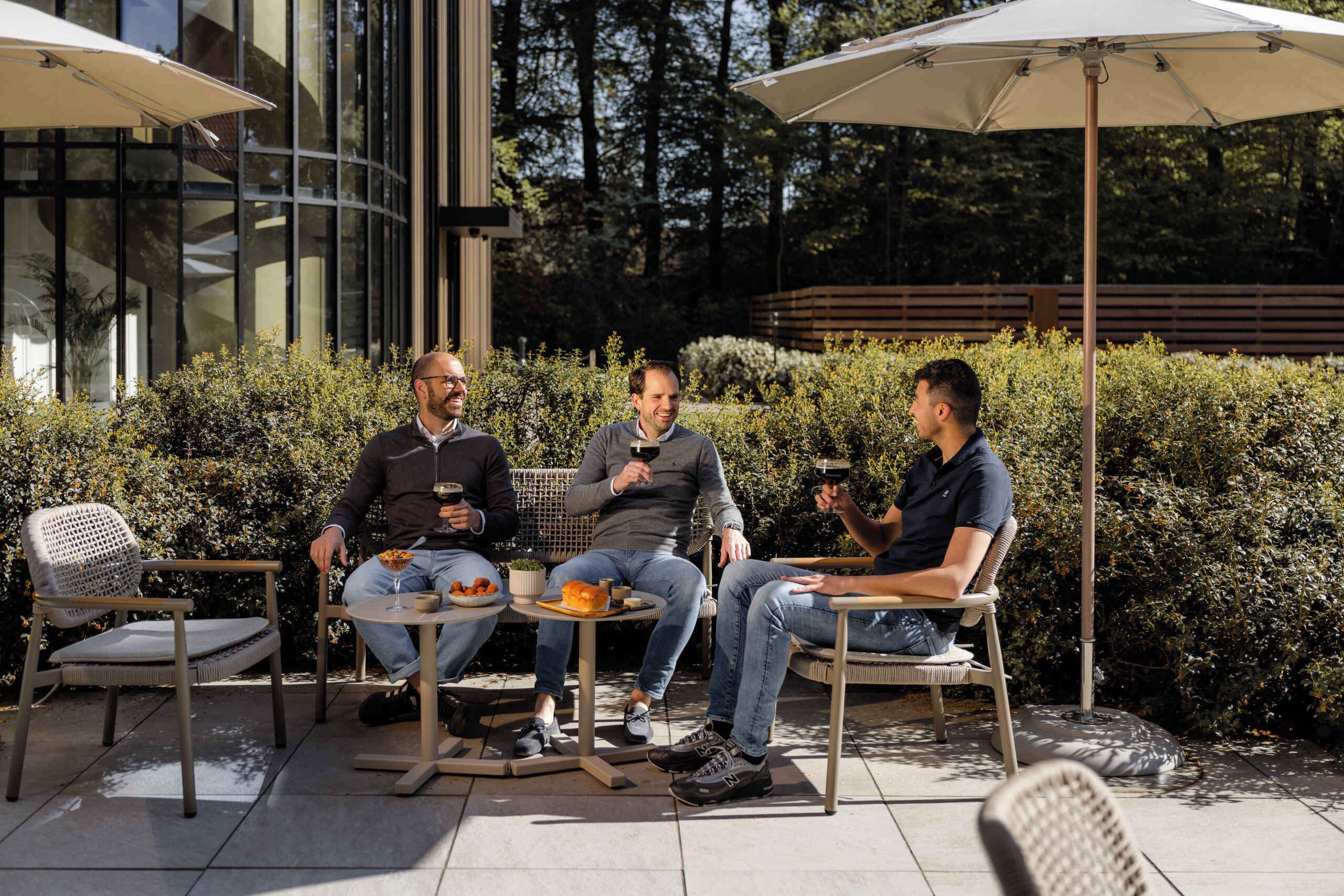 Three men seated at an outdoor table with umbrellas, surrounded by greenery and a modern building.