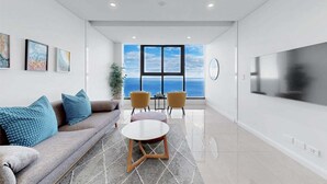 Una sala de estar moderna con un sofá, sillones, una mesa de centro y un televisor grande. Se ve una ventana con vista al océano.