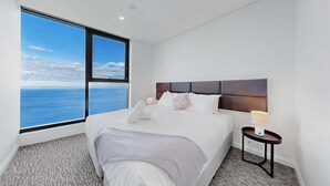 Habitación de hotel con un ventanal grande que ofrece vistas al mar, una cama bien hecha y una mesita de noche pequeña con una lámpara.
