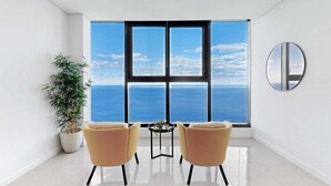 Habitación moderna con ventana grande que ofrece vista al mar, dos sillas, una mesita y una planta en maceta.