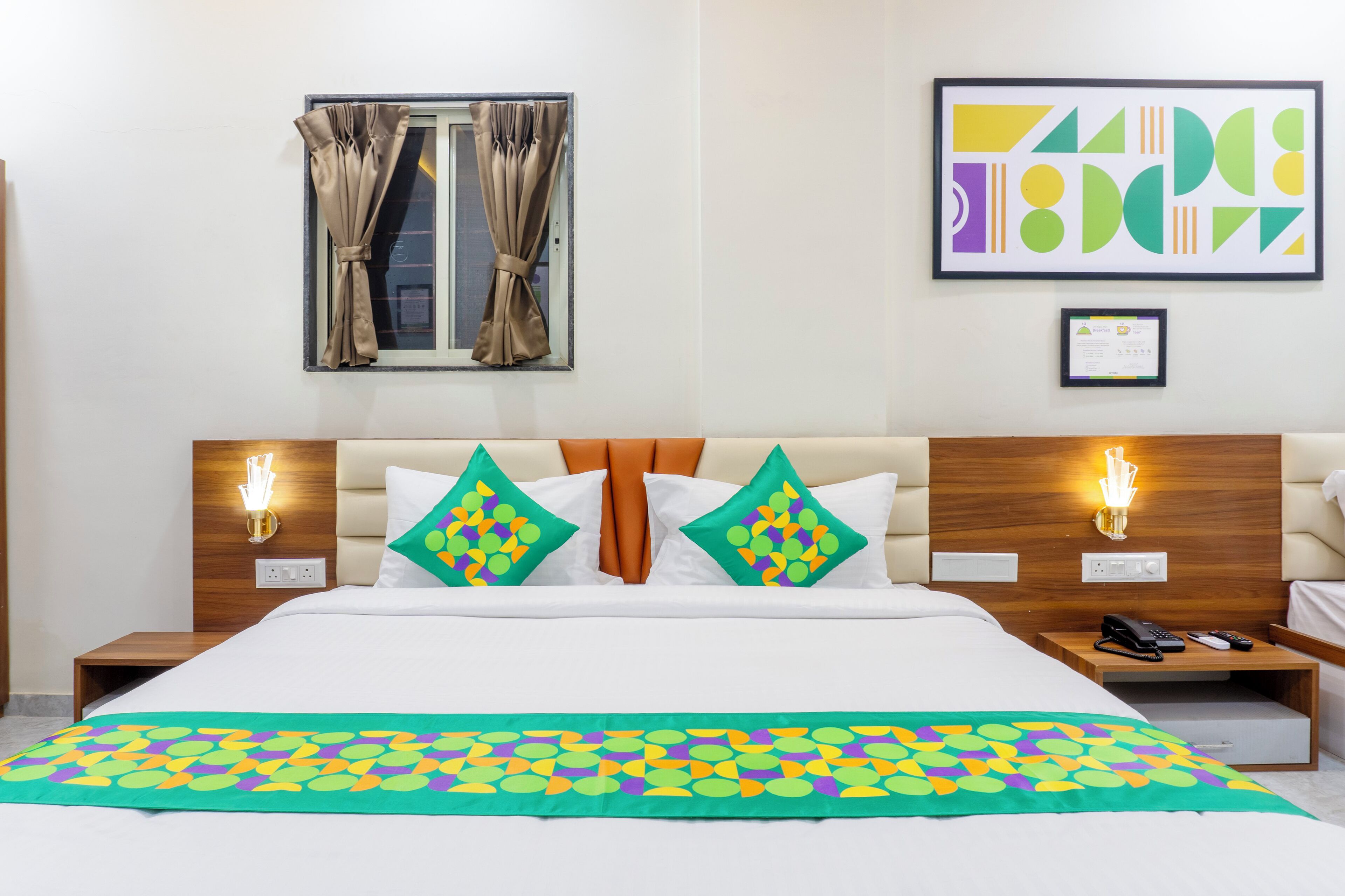 A hotel room with a large bed, two bedside tables with lamps, a window with curtains, and a framed abstract artwork on the wall.