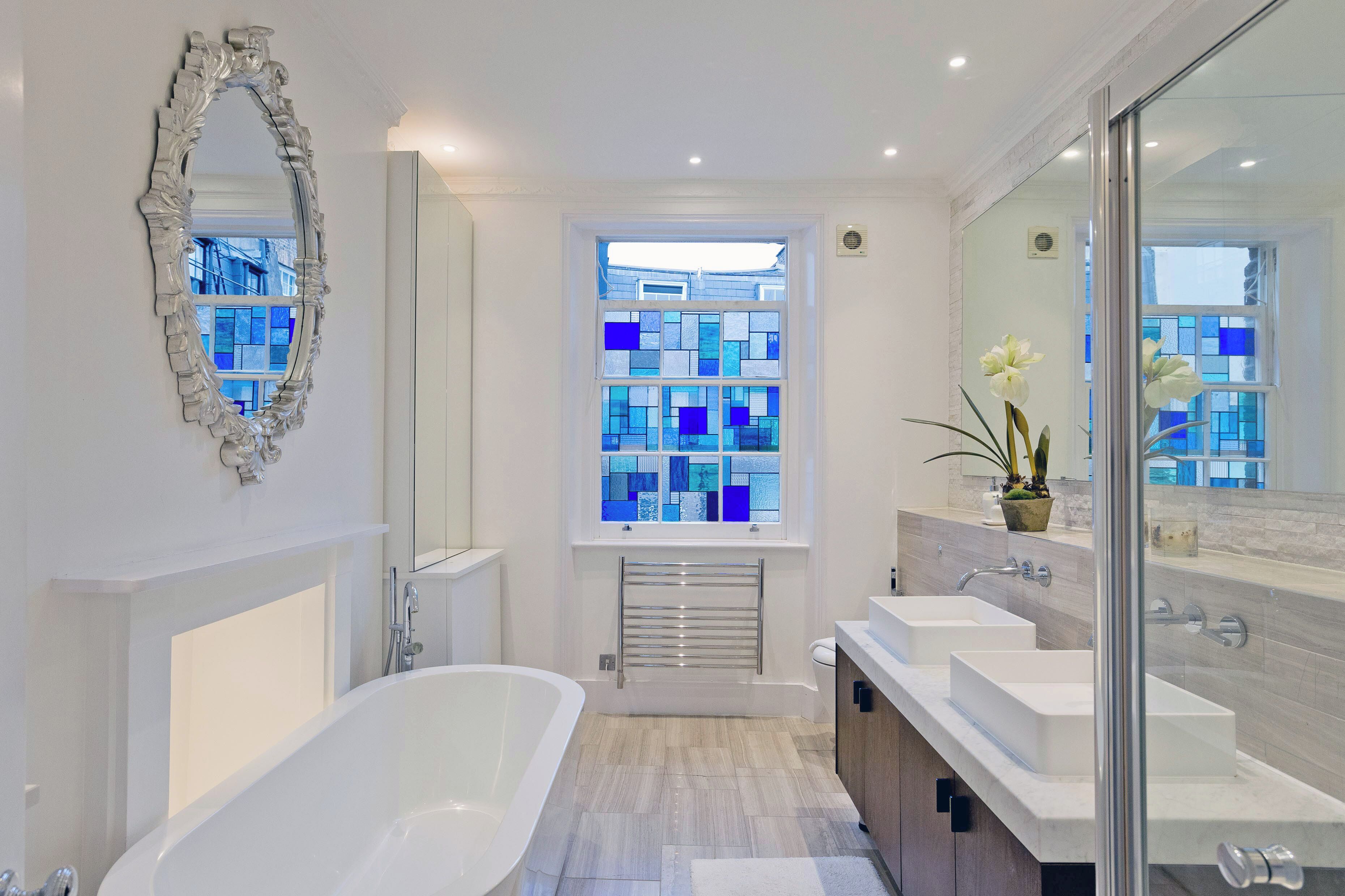 Une salle de bain moderne avec une baignoire sur pieds, un grand miroir et une fenêtre avec du verre teinté en bleu.