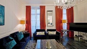 Een woonkamer met een donkerblauwe bank, twee fauteuils, een salontafel, een televisie en een kroonluchter.
